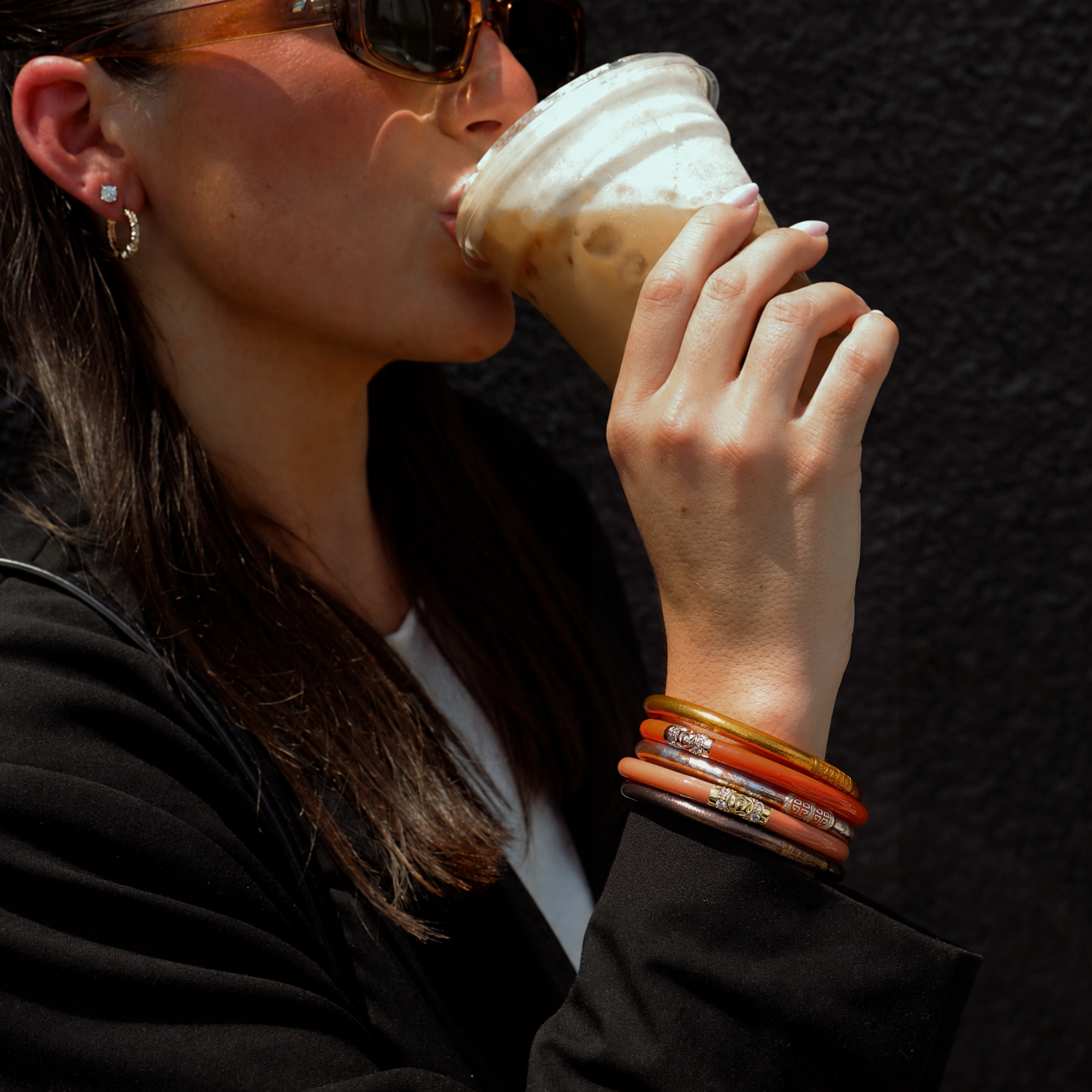 A close-up lifestyle shot of a woman wearing stacked bangles, sunglasses, and small hoop earrings while drinking from an iced coffee. The bangles feature a mix of gold, orange, and brown tones with some silver engraved details.
