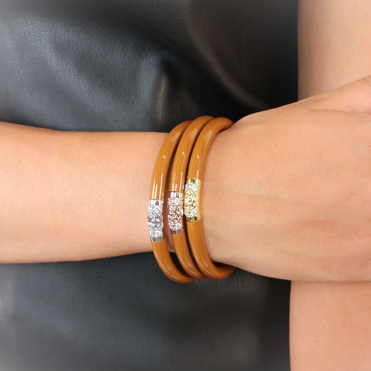 Three stacked bangle bracelets on a person's wrist, featuring a glossy tan finish with silver and gold-toned filigree center bands, set against a dark leather background.