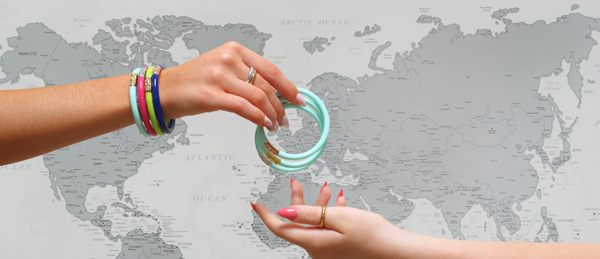 Hands holding and exchanging colorful bangle bracelets against a detailed world map background with turquoise pink blue green and gold bands