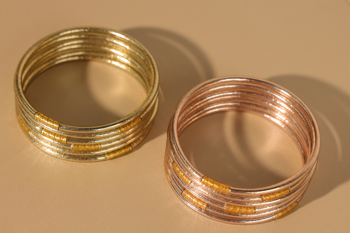 Two stacks of slim gold and rose gold bangle bracelets on a warm beige surface with soft shadows