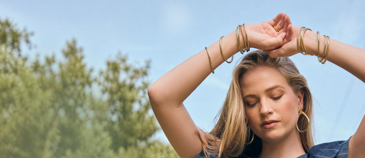 Woman outdoors with arms raised, wearing stacked gold bangle bracelets and hoop earrings against a blue sky and leafy trees