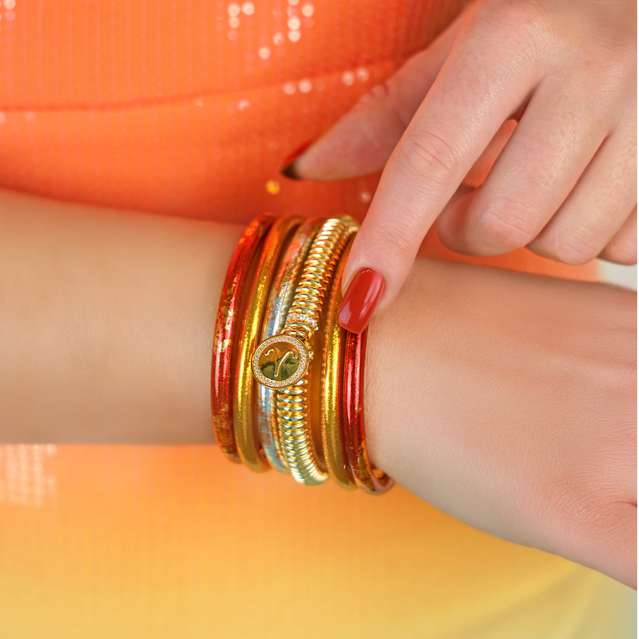 Close-up of a woman's arm wearing red and gold bangles against an orange sequined fabric background.