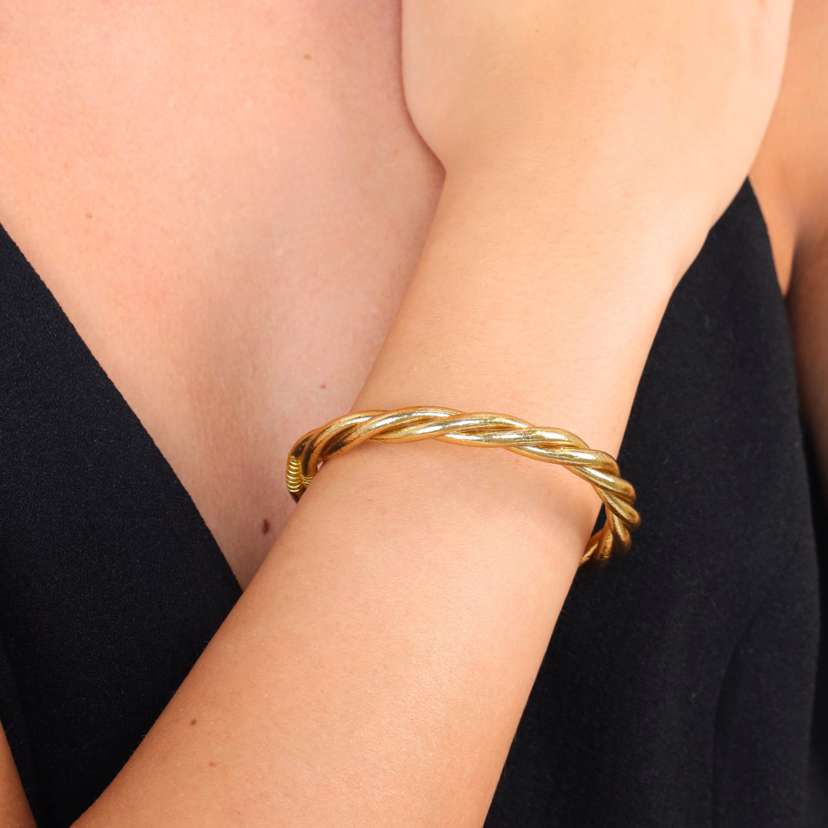 A close-up of a person's wrist wearing a twisted gold bangle bracelet, with a black top visible in the background.