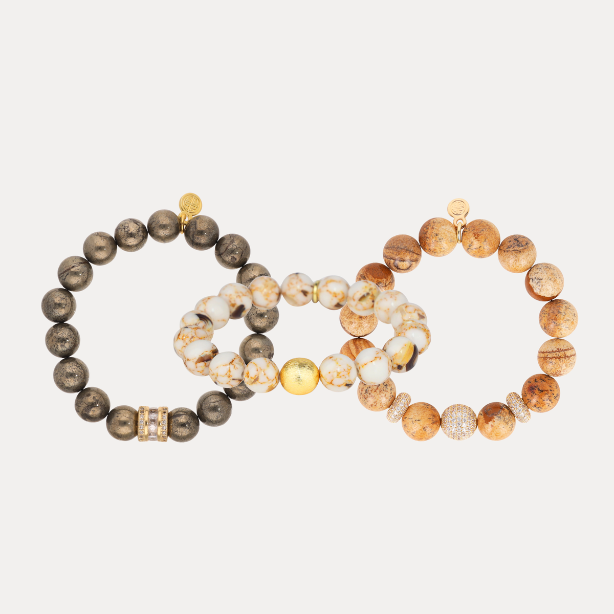 Three beaded stretch bracelets arranged on a light gray background. One bracelet is dark brown with metallic gold accents, one is cream and white marbled with gold accents, and one is tan and brown marbled with clear crystal-like beads.