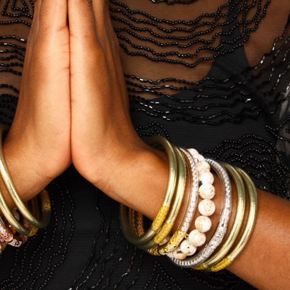 A close-up of a person's hands pressed together in a prayer pose, showcasing a stack of bohemian-style bangles on the wrists. The jewelry features gold, silver, white pearl-like beads, and textured metallic elements, worn over a black beaded lace top.