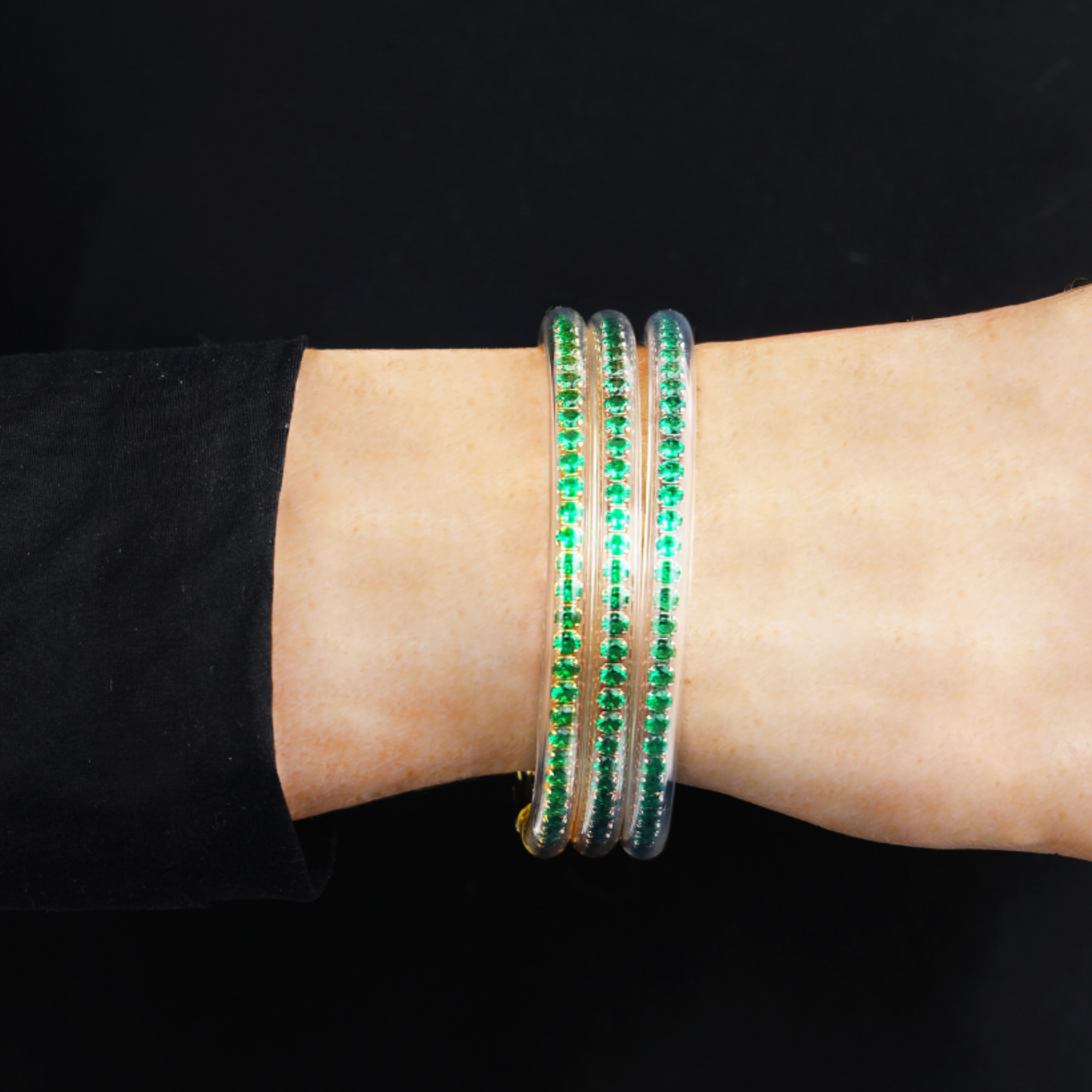 A close-up of a person's wrist wearing three slim clear bangles adorned with rows of green stones, set against a solid black background.