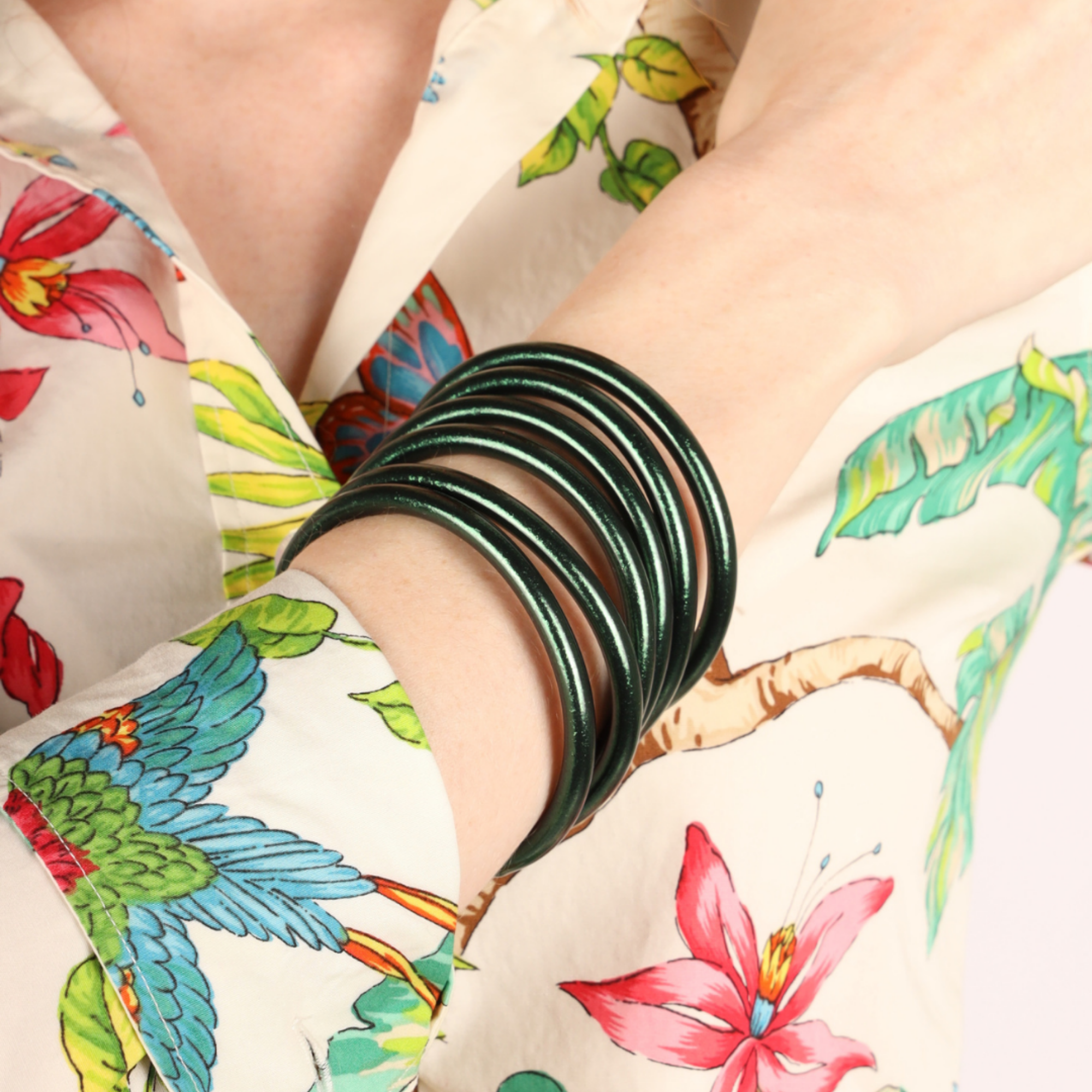 A close-up of a person's wrist wearing a stack of dark green metallic bangle bracelets, with a colorful tropical floral print shirt visible in the background.