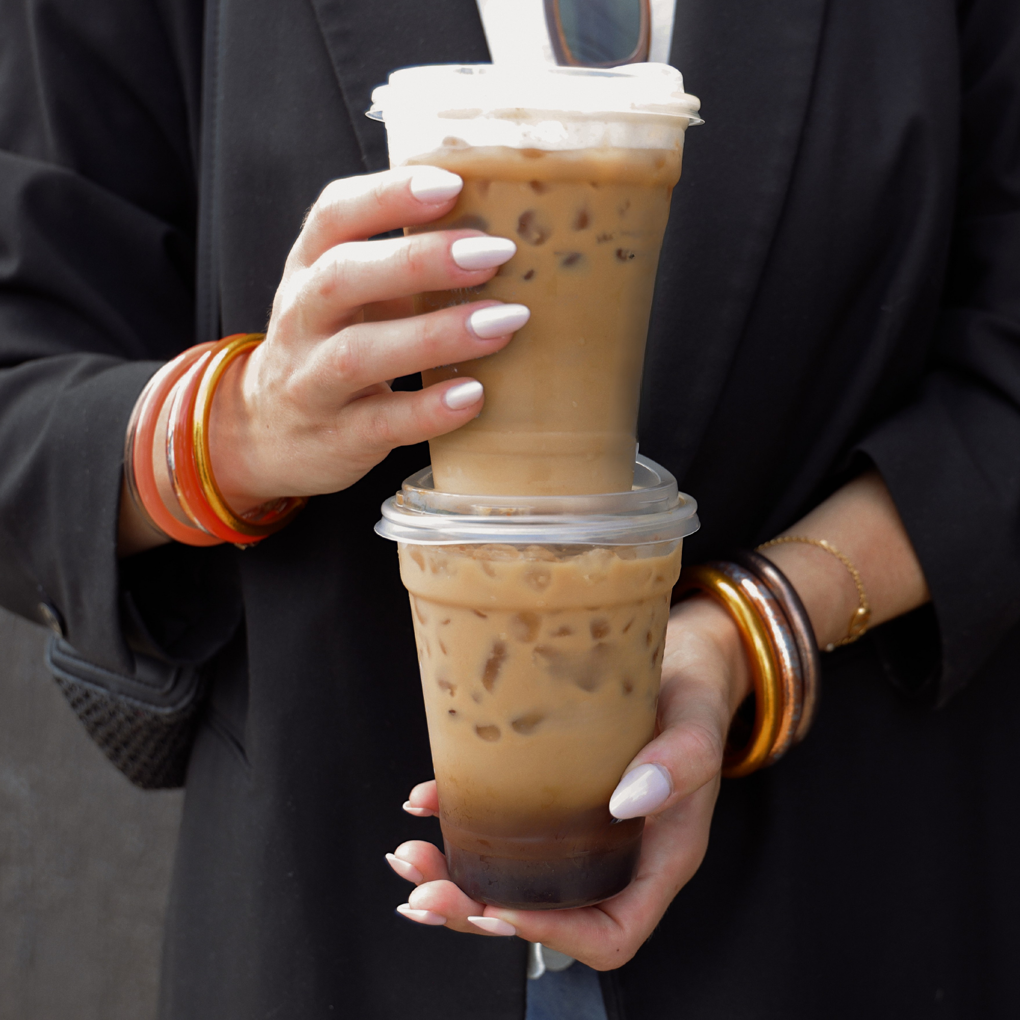 A person in a black blazer holds two iced coffee drinks stacked vertically in clear plastic cups with white lids. The person has white manicured nails and gold and orange bangles.