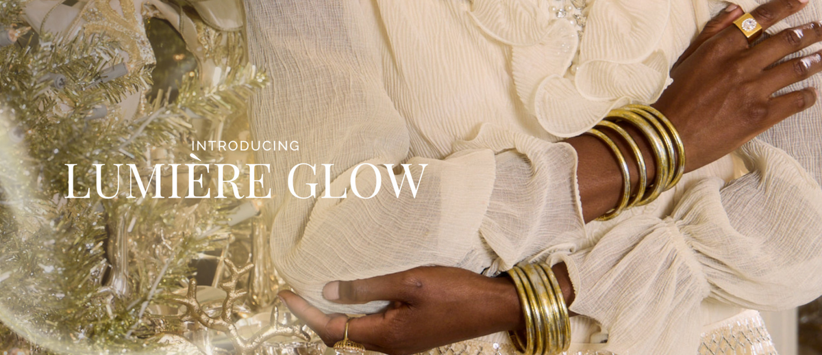 Woman in flowing cream clothing wearing stacked gold bangle bracelets beside a shimmering floral arrangement with Introducing Lumiere Glow text