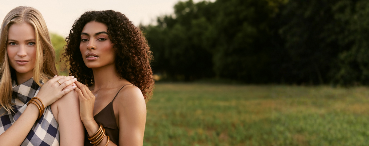 Two women standing outdoors in a grassy field with trees in the background. The woman on the left wears a blue-and-white check shirt and gold bracelets, while the woman on the right wears a brown spaghetti-strap top and stacked gold bracelets.