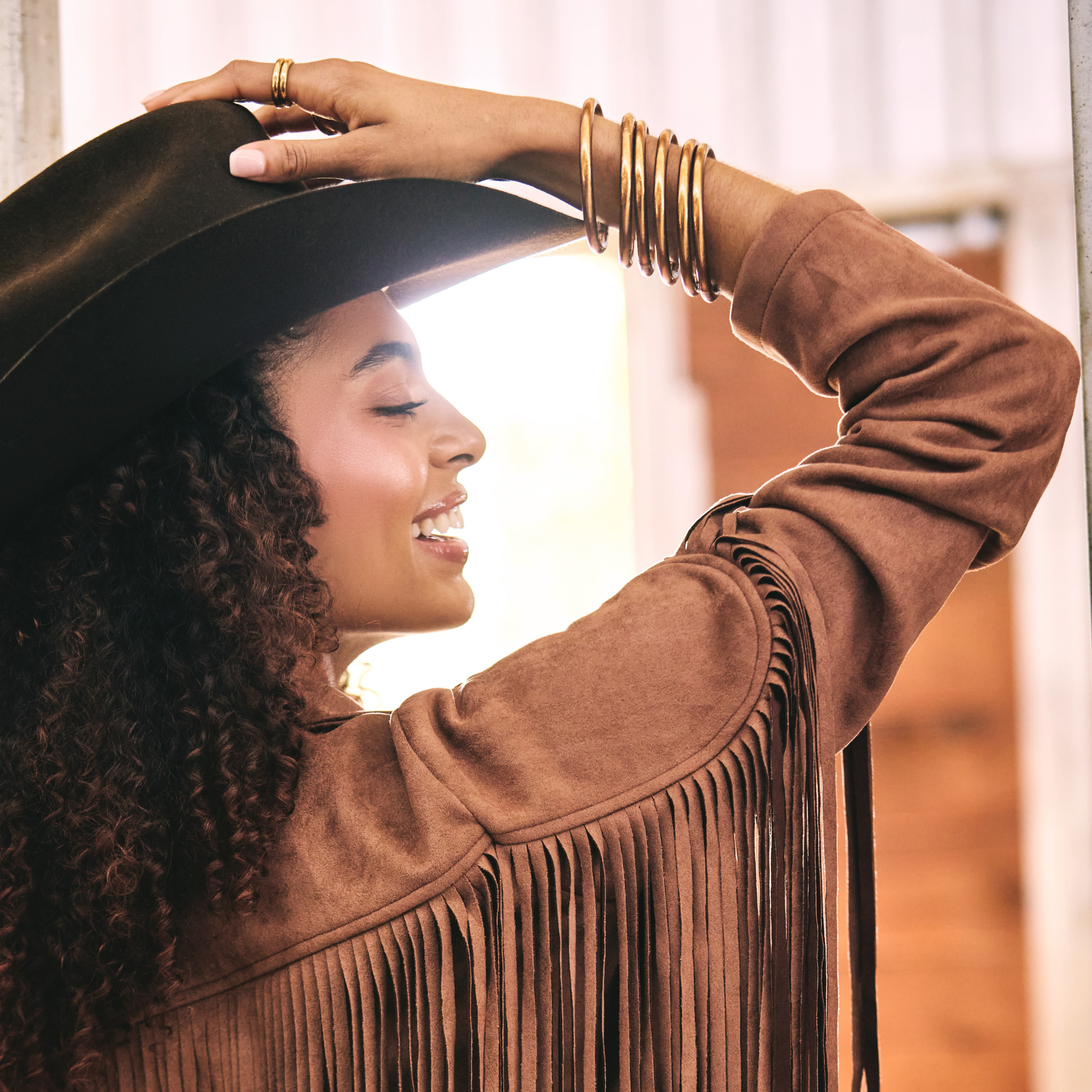 A woman in profile wearing a brown suede fringe jacket, a black wide-brim hat, and multiple gold bangles, with a bright window in the background.